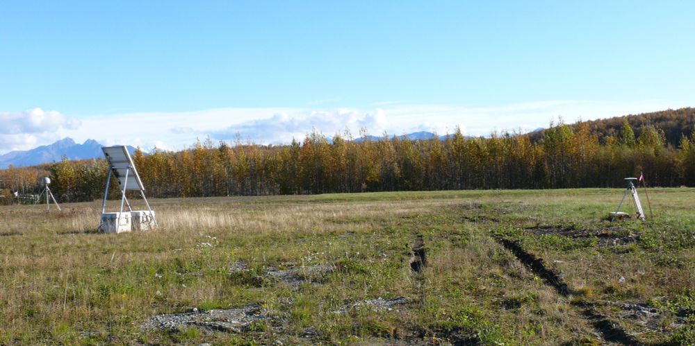 A grassy field is edged with orange and gold trees with a blue sky overhead.  Mountains peak out of clouds behind the trees.  On the left side of a field, a welded GNSS antenna monument with a gray radome (with antenna inside) stands near a solar panel array with two gray boxes holding electronics and batteries.  On the right side of the field, a yellow tripod set up over a campaign benchmark holds up a white GNSS antenna.  A yellow box with a receiver and a battery sits near the base of one of the tripod legs.