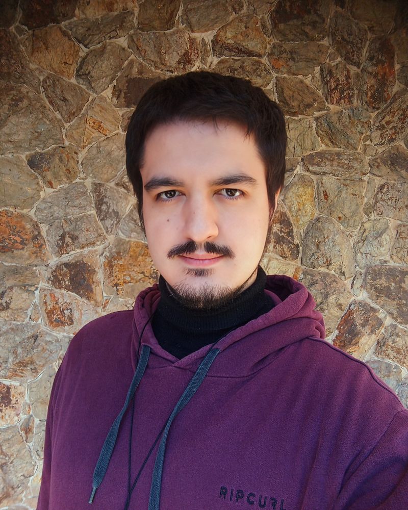 Eu, homem branco, alto, de cabelo curto escuro, olhos escuros, bigode falhado no meio, barba curta, olhando pra câmera e sorrindo de boca fechada. Estou vestindo um moletom bordô, e por baixo, uma blusa preta de gola alta. Ao fundo, uma parede de pedras, em tons de marrom claro.