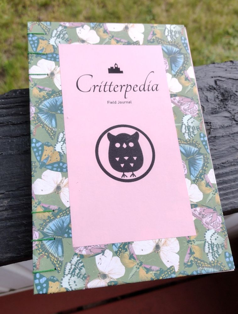 A journal covered in butterfly print cardstock. I'm the center is a pink panel and the text Critterpedia Field Journal. A small icon of a city skyline is at the top to indicate which game it's for, and a large owl icon is a bit below center below the text