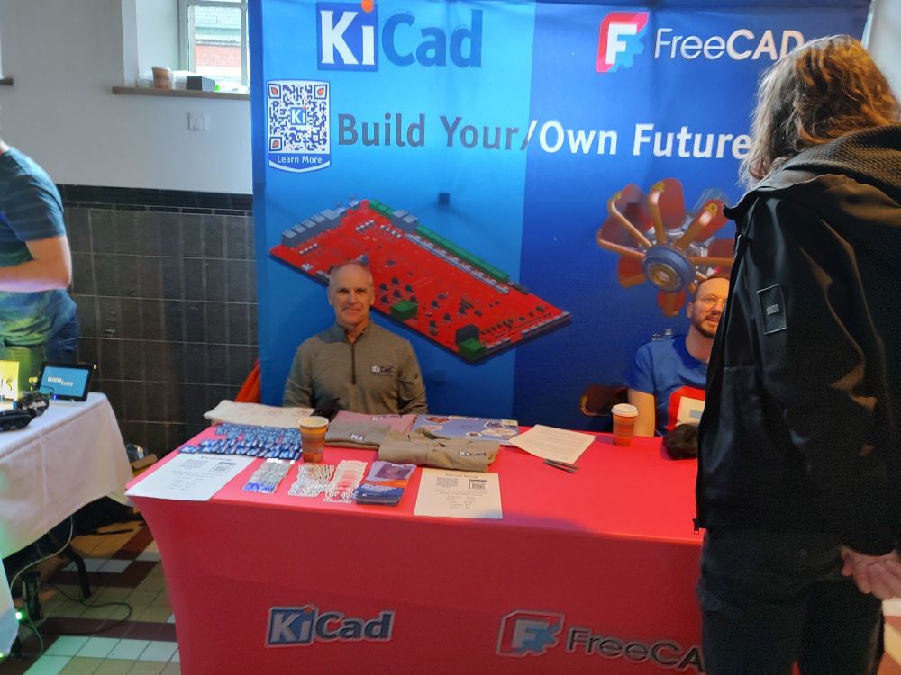 Wayne Stambaugh sitting behind a table with a freeCAD and KiCad banner in the background