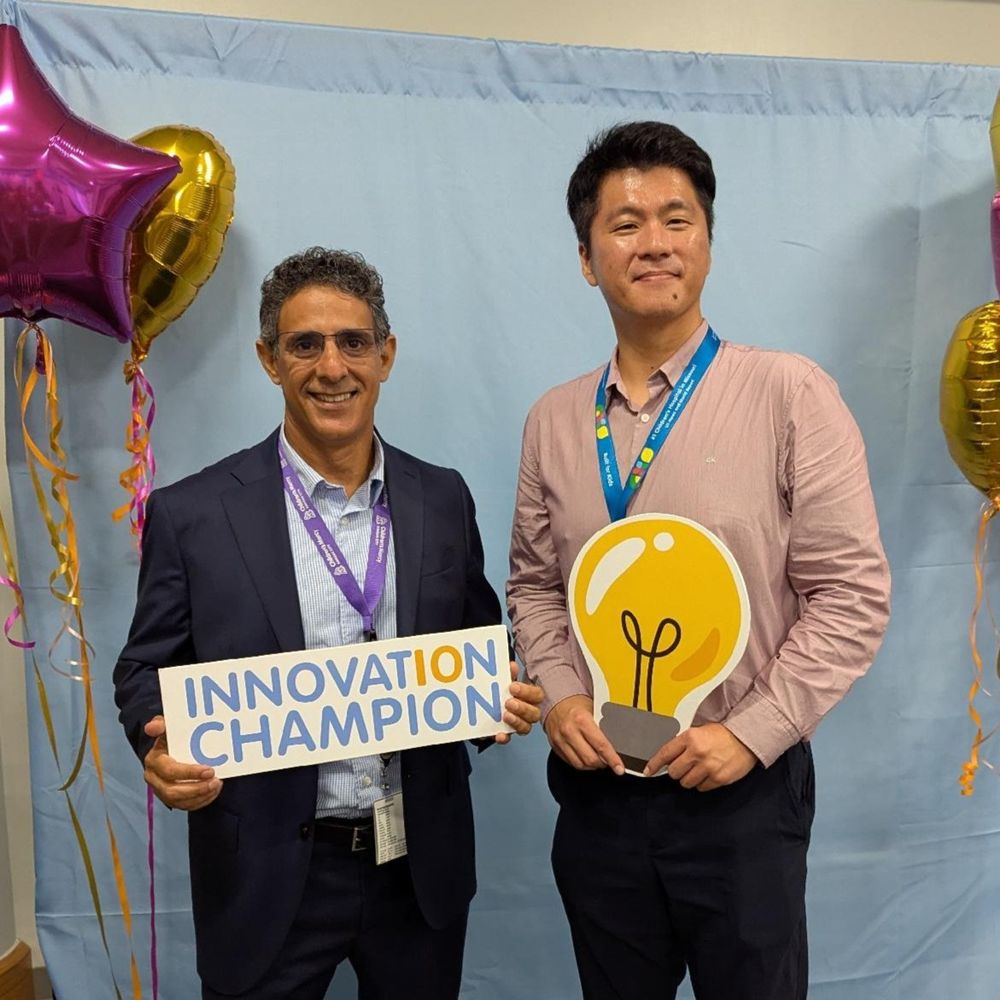 Two individuals at a corporate event, holding signs that read "INNOVATION CHAMPIONS" and a light bulb graphic, standing in front of a blue backdrop by gold and pink star-shaped balloons.