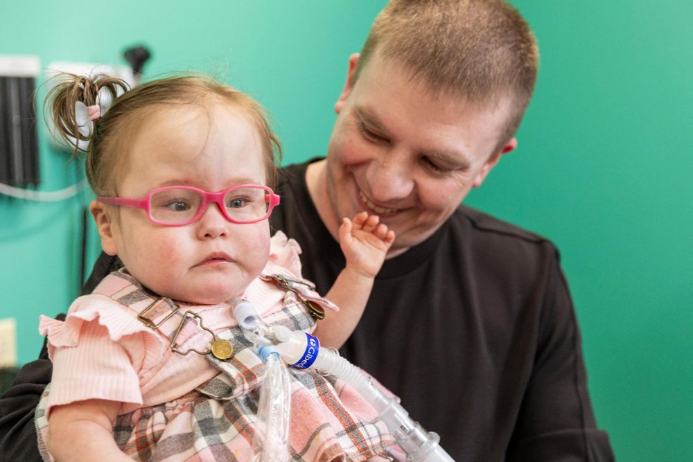 A person smiling at a young child with glasses, who is being supported in their arms. The child is equipped with medical devices.
