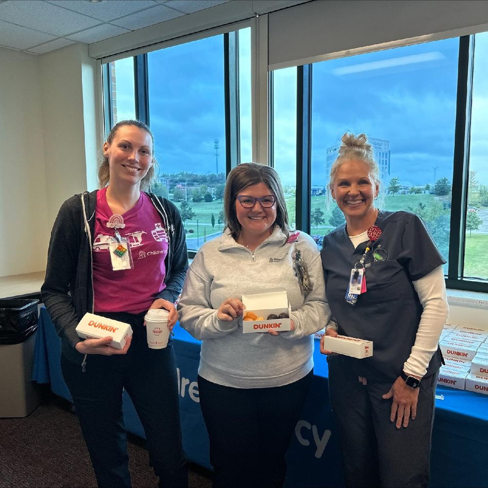 Three health care professionals holding Dunkin' gift boxes, standing in a hospital room with a view of the cityscape through the window behind them.