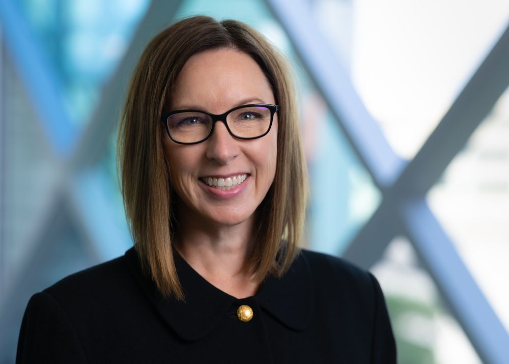 Portrait of a smiling person wearing glasses and a black blazer, standing in front of a blurred architectural background.
