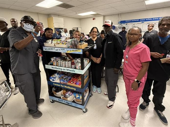 A group of hospital staff members gathered around a snack cart labeled "Wellbeing on Wheels" provided by the Center for Wellbeing, showing various healthy snacks and beverages.