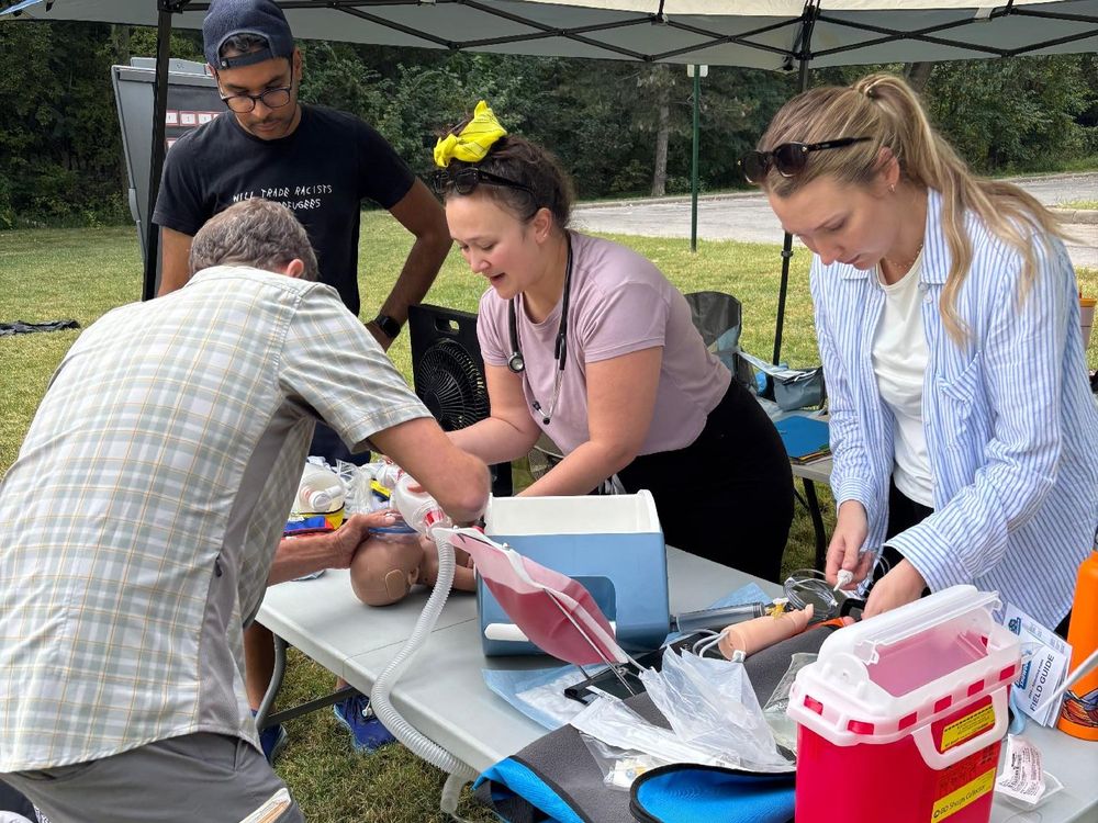 Three individuals are engaged in a medical training session outdoors under a canopy. They are focused on applying medical techniques on a mannequin arm, surrounded by medical supplies.