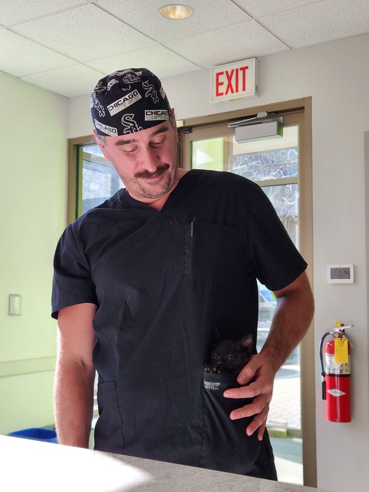 A man in black scrubs and a White Sox scrub cap with a tiny kitten riding in his left lower front pocket.
