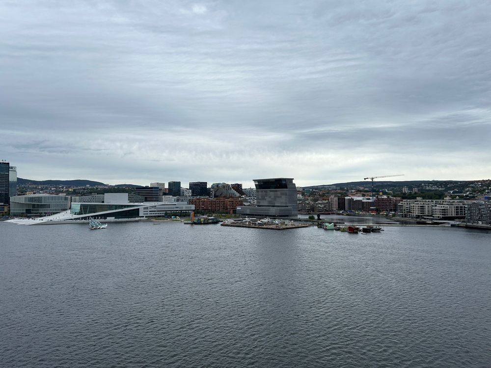 A waterfront cityscape features modern architecture, including sleek buildings and a prominent structure with an angled roof, under a cloudy sky.