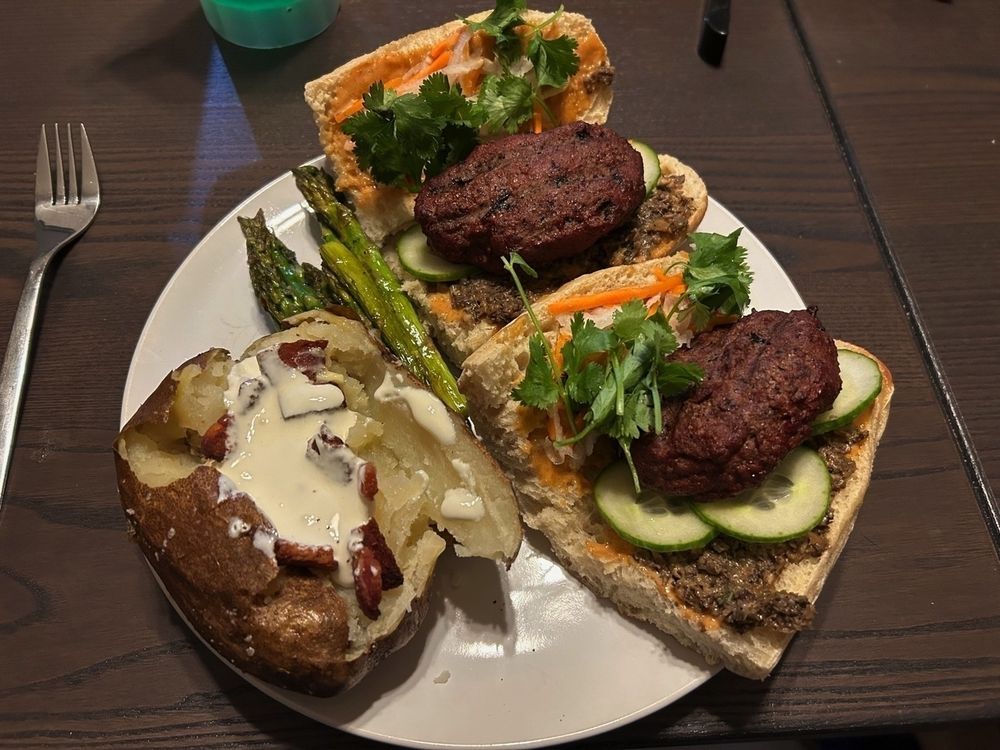 A photo of a dinner plate with two banh mi burgers open faced and a baked potato with sour cream.