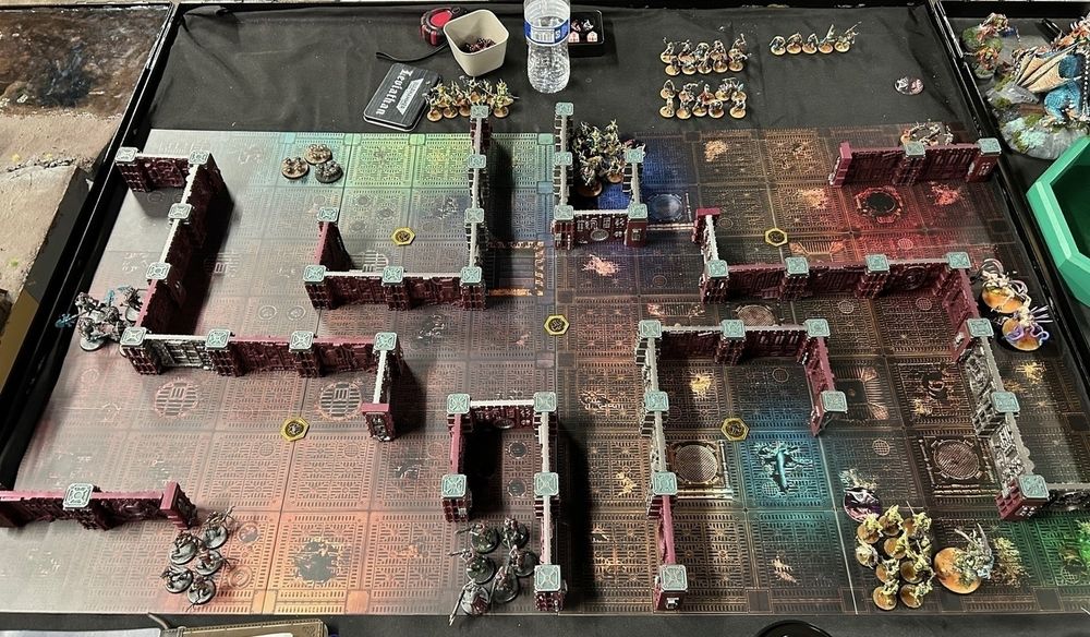 This image shows a Warhammer 40,000 tabletop game in progress, viewed from above. The battlefield is set up with reddish-brown modular walls creating a maze-like pattern of corridors and rooms across a dark gaming board. The board itself features detailed circuit-like patterns, and the walls are connected by turquoise decorative elements at their intersections. Several groups of painted miniature figures are positioned around the board, representing different armies with distinct color schemes - some darker units and some in yellower tones. A few yellow objective markers are scattered across the battlefield, and gaming accessories like a water bottle and small containers for dice or tokens can be seen at the board's edges.