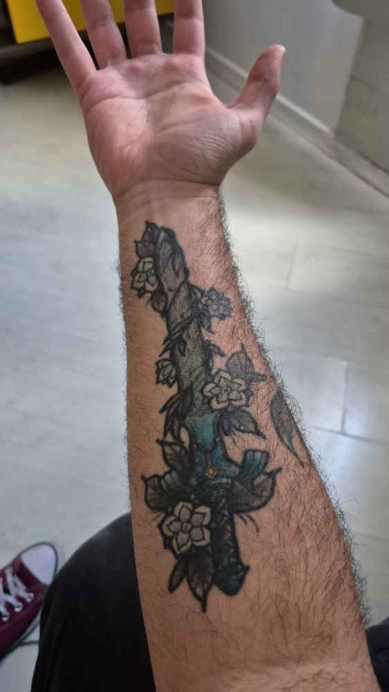 Antebraço com tatuagem old-school de uma espada azul envolta por flores
