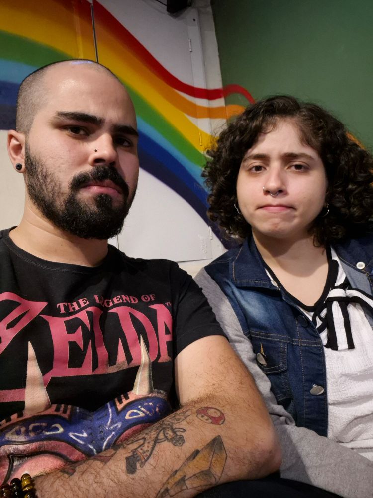 Homem careca e barba, com camisa de Zelda, sentado ao lado de uma mulher de camisa branca e jaqueta jeans. Ela tem cachinhos pretos, muitos brincos e tem um rosto que me faz querer rebolar lentinho pra ela, te amo bb 🫶🏼