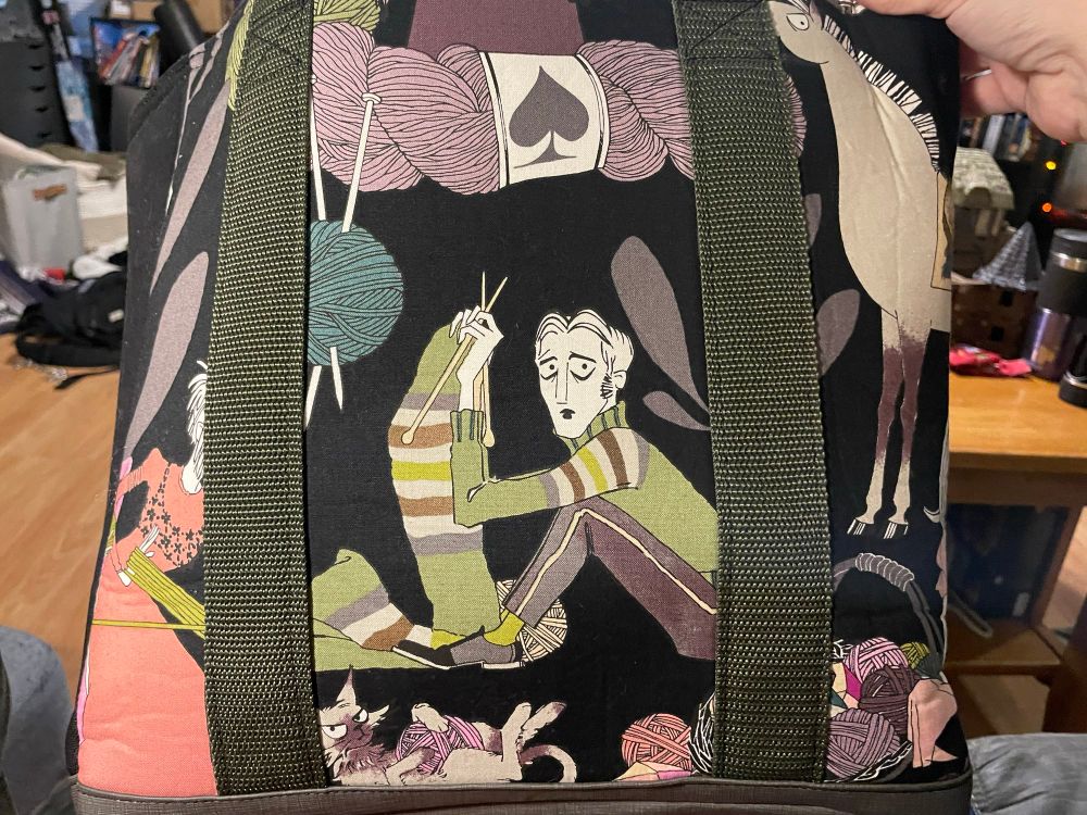 Central on this tote is the image of a young man knitting a scarf. Obscured on the left appears to be a woman helping wind yarn. A grumpy looking cat holds a ball of yarn at the bottom. There is a partial view on the right of a horse above a basket of yarn and the barest glimmer of another figure at the bottom right.