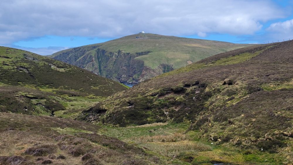 Looking through a heather-clad gully towards the distant conical hill of Saxa Vord on the island of Unst. There is a white radar dome on top of the hill