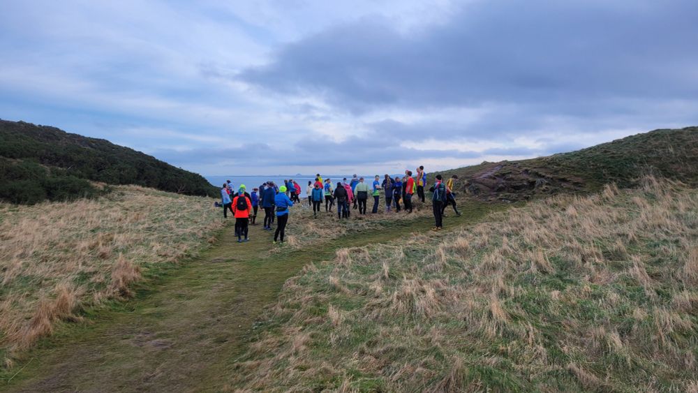 A group of orienteering in brightly colored clothing gathered at the course start in an area of rough grass and scrub. The sea is visible in the distance 