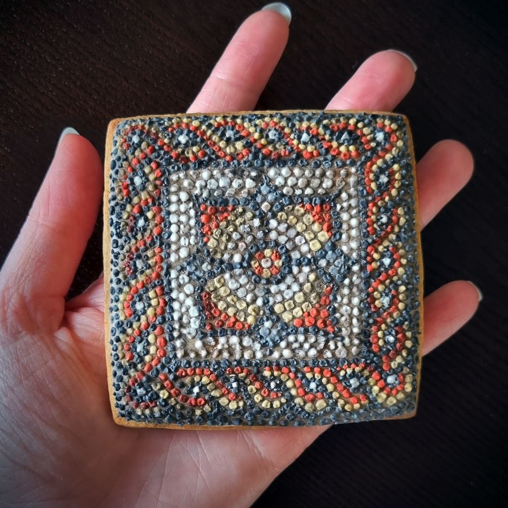 A square biscuit decorated with tiny rough squares of royal icing, creating a mosaic effect. In the centre of the design is a four-leaf flower in shades of red, yellow, and cream. Around the edge of the design is an intertwining strand motif, in shades of red, yellow, and grey. The surface of the biscuit is dirty to create the effect of an excavated mosaic.