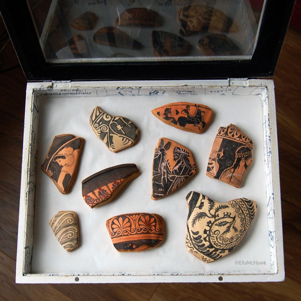 A set of nine unevenly shaped biscuits arranged in a glass-lidded box. Each biscuit is curved and made to look like a fragent of ancient pottery. The base icing is in shades of orange, peach, and cream. Figures, swirls, repeating leaf patterns, and horses are painted onto the icing surface in black. The biscuits are aged with scratch marks and cracks. 