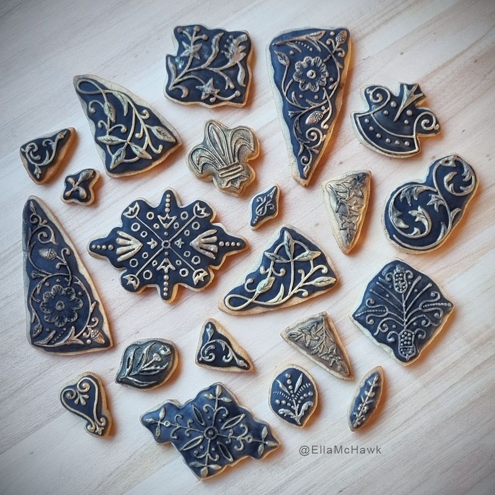 A collection of iced biscuits designed to look like decorative book binding tools. The biscuits are all different shapes and sizes: some are triangular, designed to fit snugly in the corner of a book; others are square, oval, or diamond shaped. Most of the biscuits have a patchy dark background. The designs piped onto the biscuit surfaces feature interweaving curled lines, leaves, spirals, and decorative dots. These raised, 3D designs are painted with metallic lustre dust in different shades of gold, silver, and bronze to make the biscuits look like metal stamps. The biscuits are brushed with dark powder so they appear dirty and aged.