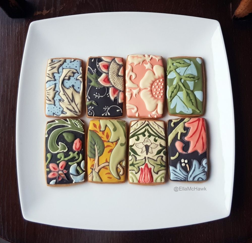 A set of eight rectangular cookies arranged on a square white plate against a dark wooden background. Each cookie is inspired by a wallpaper design by Morris & Co. The patterns represented feature different floral and leaf shapes in shades of baby blue, soft pink, leafy green, mustard yellow, cream, and dark grey.