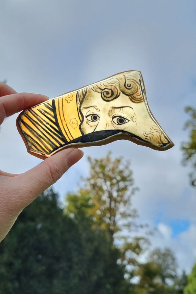 A hand holds up a biscuit with uneven edges against a cloudy sky. The biscuit has an egg-yellow background decorated with strong black lines. A man with curled hair and striking eyes features in the foreground of the design. Only his eyes, eyebrows, and forehead are visible; a jagged break line at the bottom of the biscuit shows where the design would continue.