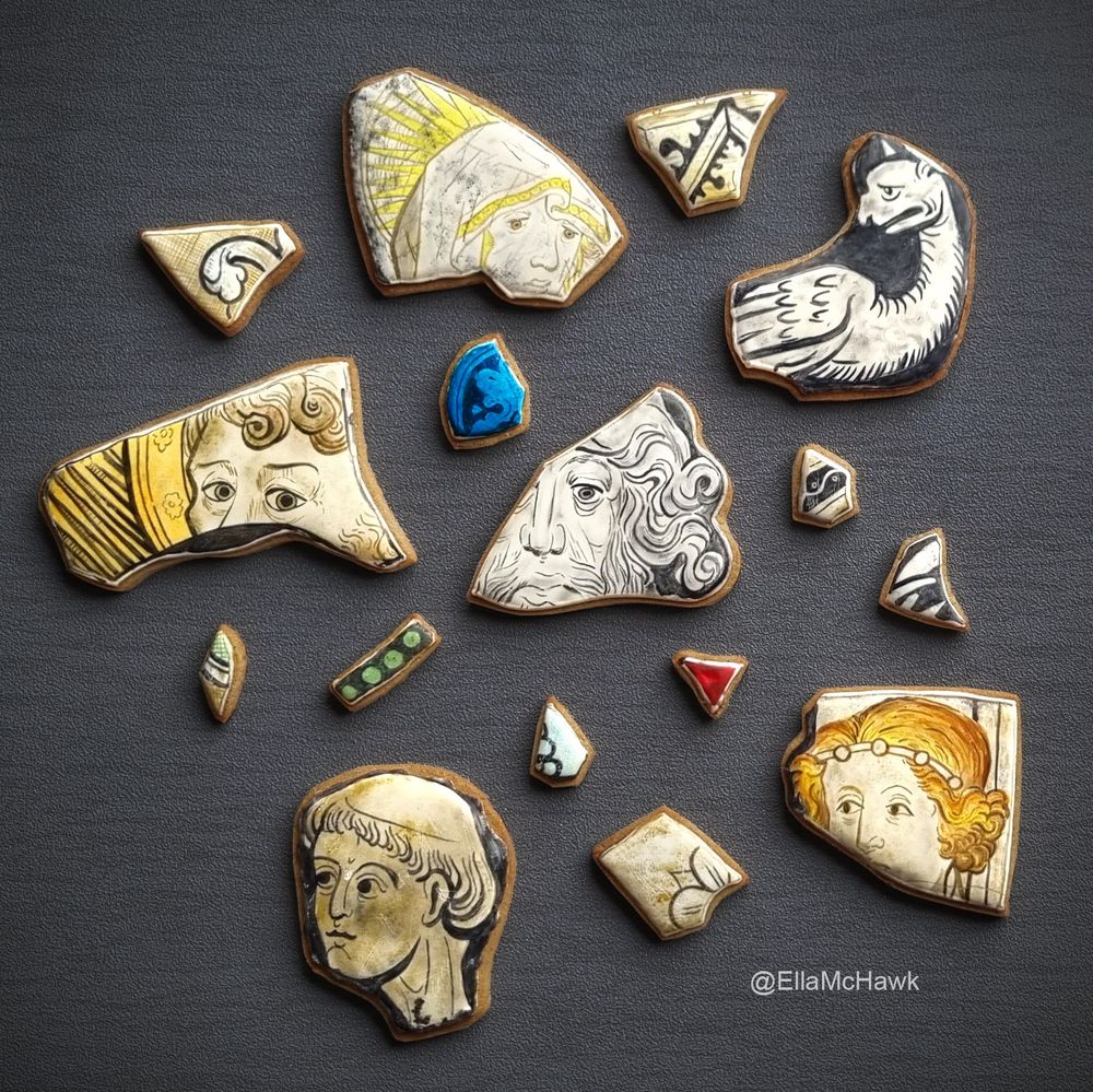 A collection of biscuits made to look like stained glass window fragments. There are six larger biscuits featuring different faces: a woman looking downwards; a griffin-like creature with wings and a long neck; an old man with long facial hair; a woman with golden hair and a simple tiara; a bald man painted in monochrome; a man with curled hair and striking eyes. The larger biscuits are surrounded by a scattering of smaller fragments. There is a jewel-red fragment in a triangular shape, a small green rectangle, and a bright blue semi-circular shard painted with a leaf pattern. Other small biscuits have bold black-and-white patterns made up of lines, circles, and floral shapes. Many of the biscuits are made to look dirty and scratched.