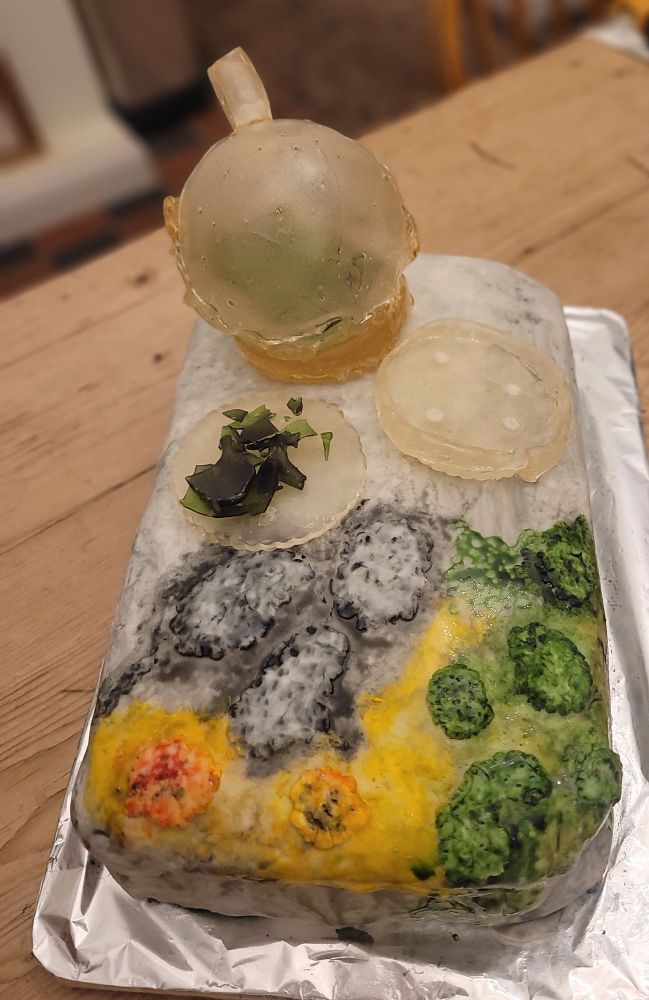 A cake decorated with icing and food colourings to look like a gravestone with lichen growing on it. There are also some laboratory glass ware items made (badly) out of isomalt sugar on the cake.