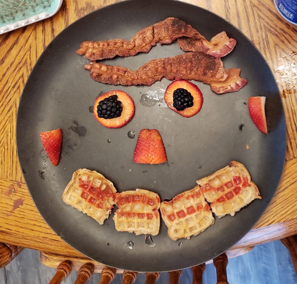 A disturbing smiley face constructed of berries (eyes, nose, and ears), mini waffles with strawberry syrup (mouth) and bacon (hair.)