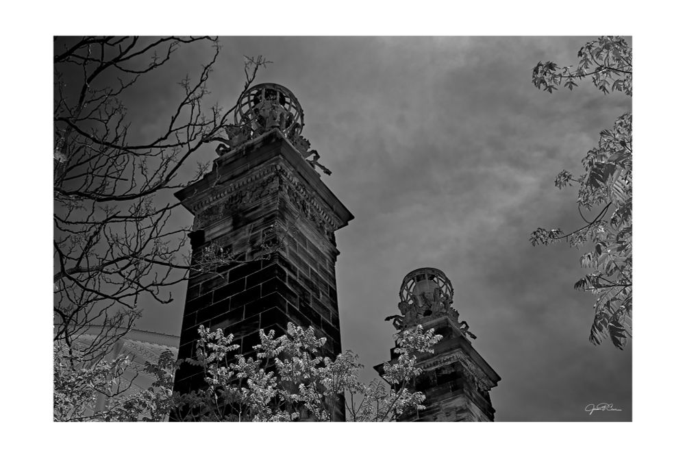 A dramatic black and white photograph of two ornate stone pillars rising above a cluster of leafy and bare tree branches. The towering pillars are crowned with classical sculptures—armillary spheres encircled by rearing horses and human figures, suggesting themes of celestial navigation or mythological symbolism. The image is shot from a low angle, enhancing the monumental feel of the architecture. The cloudy sky above swirls with texture, adding to the mysterious and almost otherworldly mood. The foliage in the foreground contrasts sharply with the stark lines and shadowed depth of the stone, creating a powerful interplay between nature and man-made form.