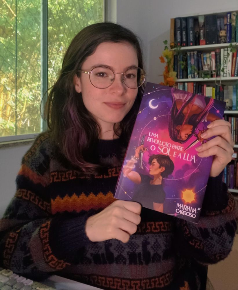 A autora, uma mulher branca de óculos e suéter, segurando seu livro Uma Revolução Entre o Sol e a Lua. 
Descrição da capa:
Uma mulher branca de cabelos bem curtos e castanhos, blusa preta, tatuagem de lua no pescoço e um feixe de magia enrolado no pulso com um bracelete. Ela segura uma adaga de lua. No meio da capa vem o título. Na parte de cima, uma mulher negra, tranças rosas e roxas, com algumas miçangas e vestindo uma blusa rosa. Ela segura uma adaga de sol. O fundo imita um céu/galáxia em tons de rosa e roxo e há circulos com estrelas, sol e lua. No canto direito inferior o nome da autora Mariana Cardoso.
