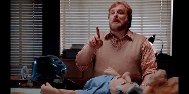 A red haired bearded man answers questions at his desk about his kids TV show “Smile Time”. Not pictured, the evil puppet puppeteering the man through a bloody hole in his back. 