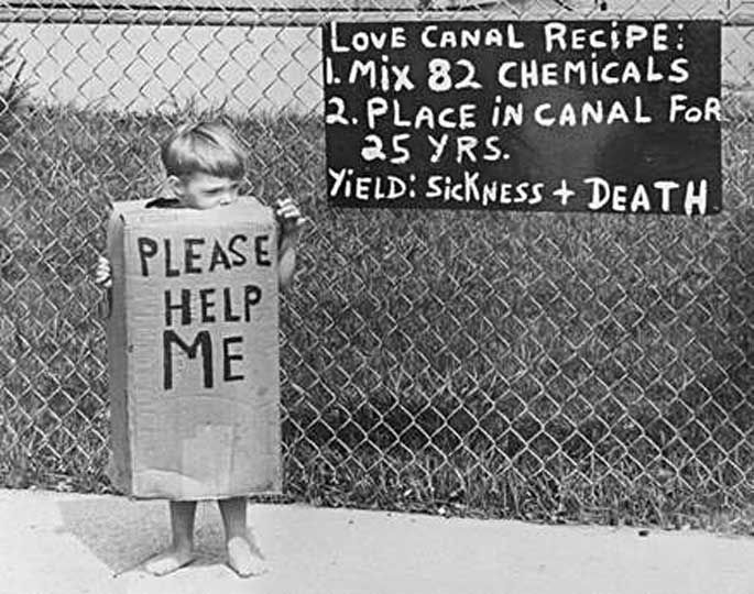 A child wears a cardboard box with the message "Please Help Me," protesting the environmental disaster at Love Canal. The sign on the fence reads: "Love Canal Recipe: 1. Mix 82 Chemicals. 2. Place in Canal for 25 Yrs. Yield: Sickness + Death."