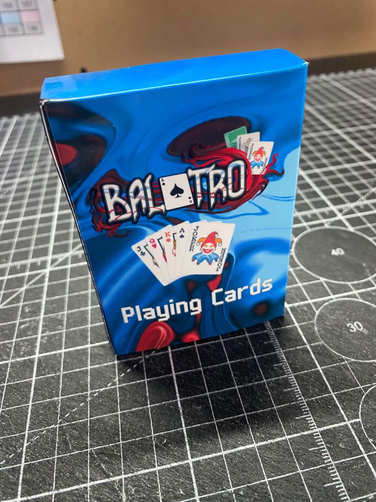A close-up image of the "Balatro" playing cards box. The design features a vibrant blue background with a swirling pattern of red and black accents. The logo "Balatro," styled with a playing card theme, is prominently displayed above an image of a fan of cards, including a Joker card with a playful design. The words "Playing Cards" are written in white at the bottom. 