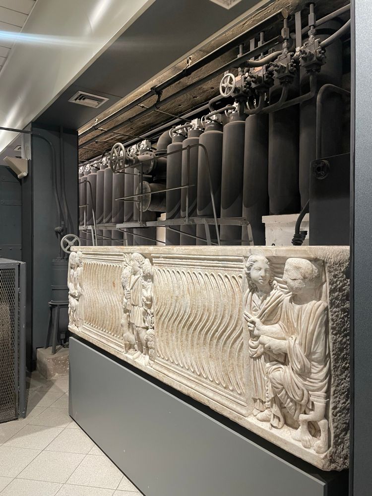 An ancient Roman sarcophagus with a wavy vertical motif in front of huge vertical black iron boiler pipes.