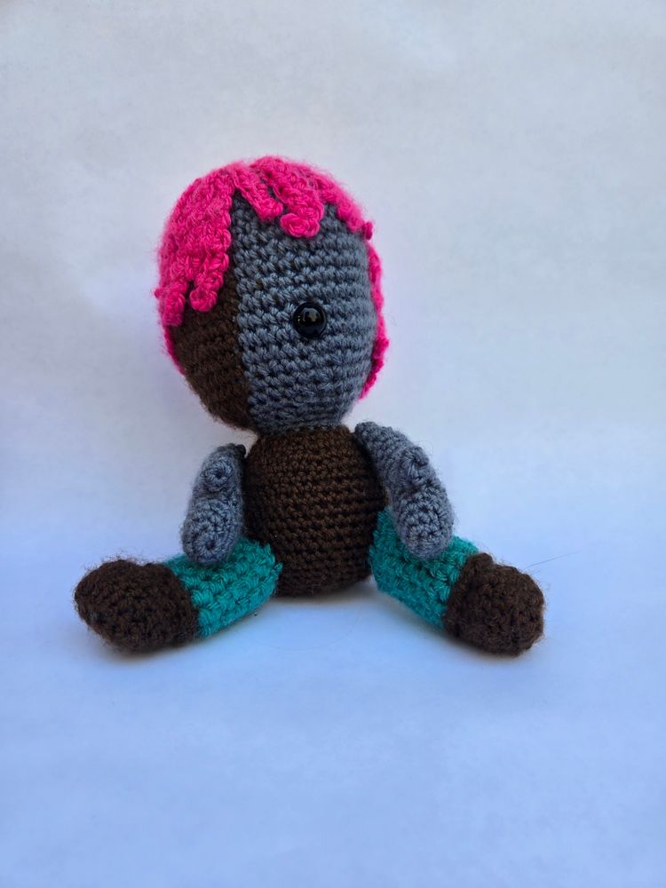A crochet doll of Gooey from d20 a crown of candy. She is purple with one eye, the other half of her face is dark brown. She has magenta hair, green pants and brown boots and body. 