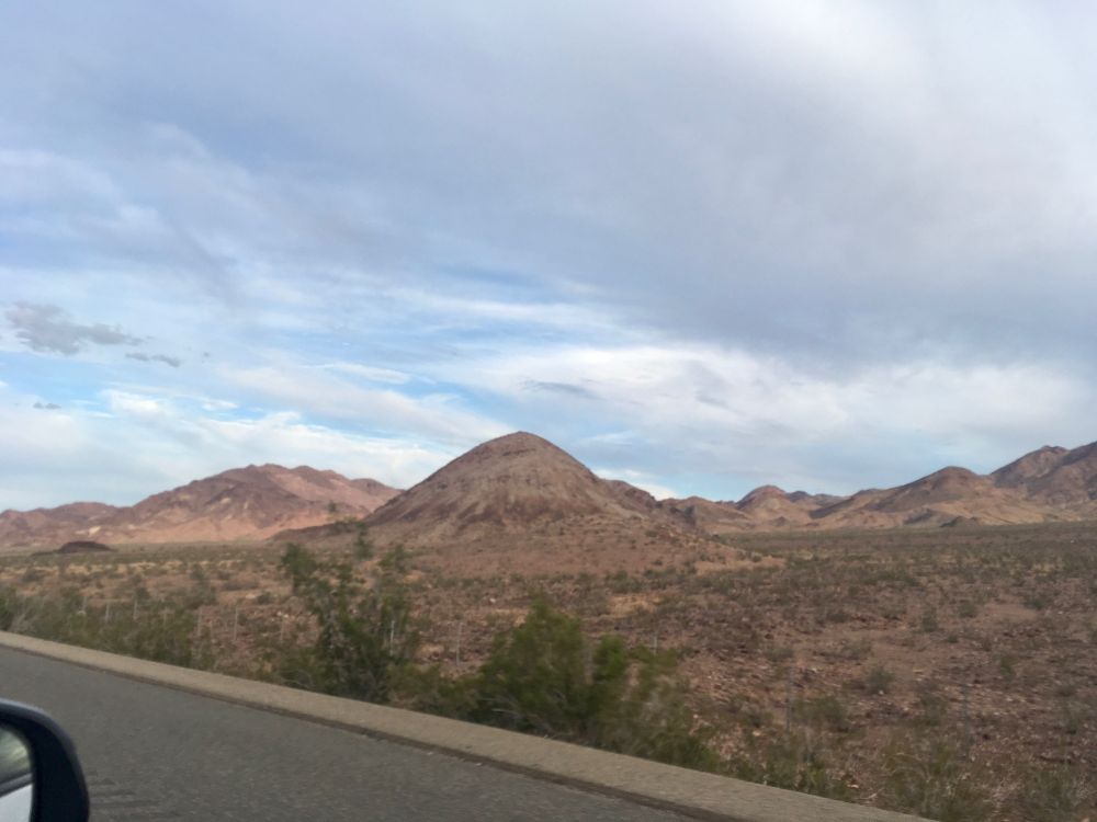A few scraggly sagebrush dot a plain of pale dirt, with round hills rising abruptly from the flat, geology of their different layers very visible with no vegetation 