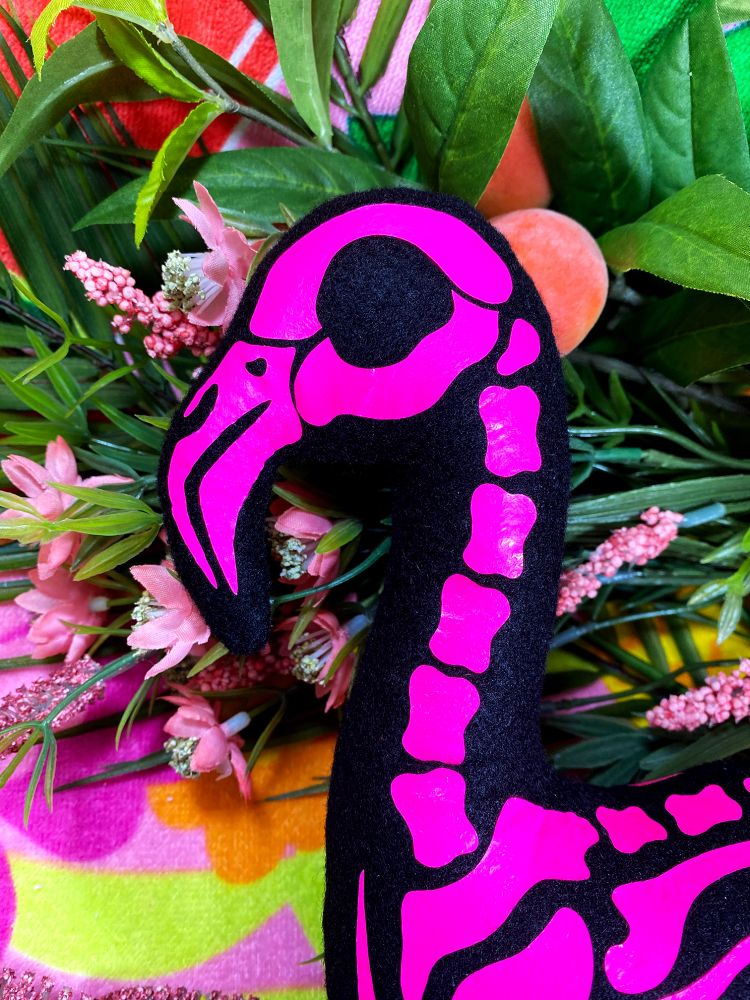 A black flamingo plush with pink vinyl applied bones. This looks like a lawn flamingo skeleton but in plushie form and bright pink! It's sitting on a tropical towel with fake tropical flowers. This is a closeup of it's head.