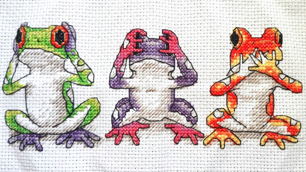 A cross-stitch depicting three colourful frogs, in the poses of ‘Hear no evil, see no evil, speak no evil’