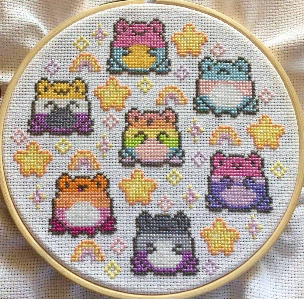 A cross-stitch showing seven little frogs, each with different Pride flag colours including: Pan, Non-Binary, Gay, Trans, Lesbian, Bisexual, and Asexual. They are surrounded by little stars, sparkles and rainbows.