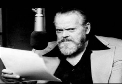 A picture of Orson Welles holding a script and sitting at a microphone 