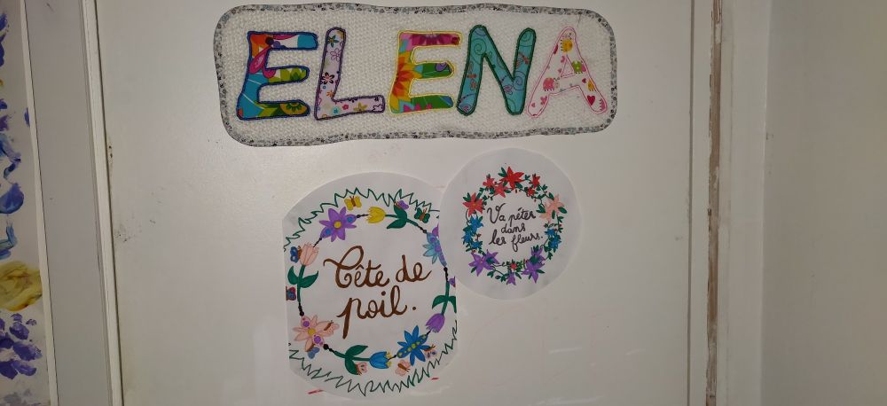 Porte de chambre avec le prénom Elena cousu en tissus multicolores sur in rectangle de serviette éponge écru, et deux dessins de sandrine Deloffre qui contiennent les textes "tête de poil" et "Va péter dans les fleurs" entourés d'une couronne de fleurs multicolores.