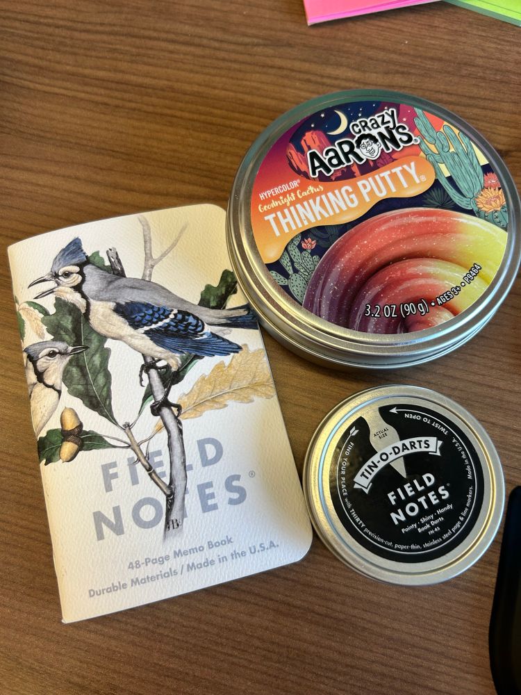 Crazy Aaron’s color changing thinking putty in a round, metal tin. Next to a Field Notes memo book with a Blue Jay illustration on the cover, next to a smaller round tin of Field Notes Brand book darts. 