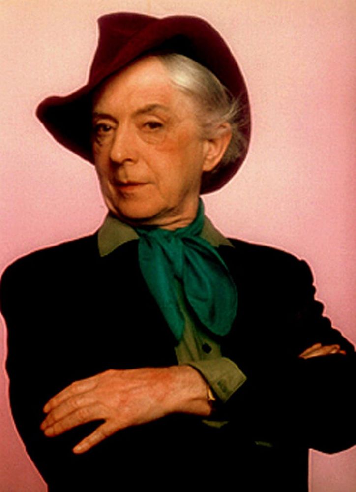 The late Quentin Crisp, striking a somewhat suspicious pose, arms crossed, looking askance at the camera, wearing a maroon fedora, a pea green shirt under a black jacket, and a jade green ascot.