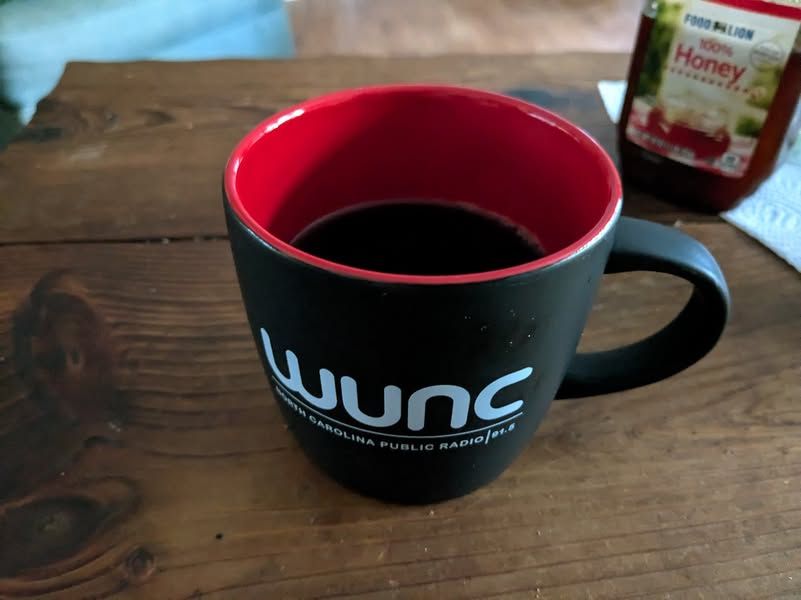 A coffee mug with black coffee in it sitting on a wooden table. The mug is matte black on the exterior, and glossy red on the interior. White printing on the mug reads "WUNC, North Carolina Public Radio, 91.5". There is a jar of honey in the background for some reason.