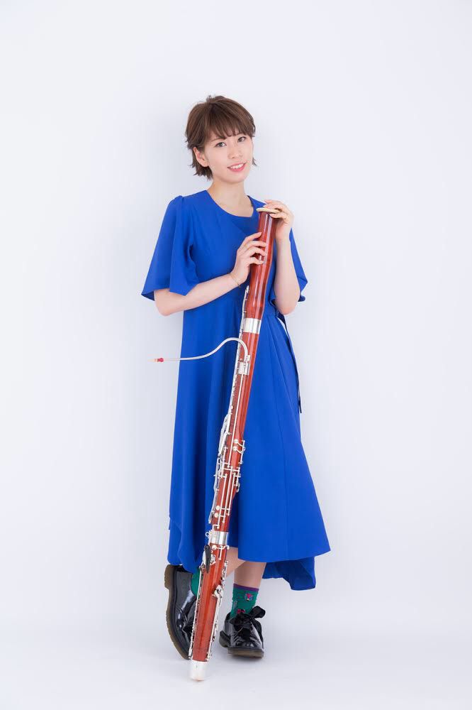 ファゴットを自分に立てかけるようにして足元に置き、硬めの笑顔をこちらに向ける河村の写真です。青いワンピースを着ています。