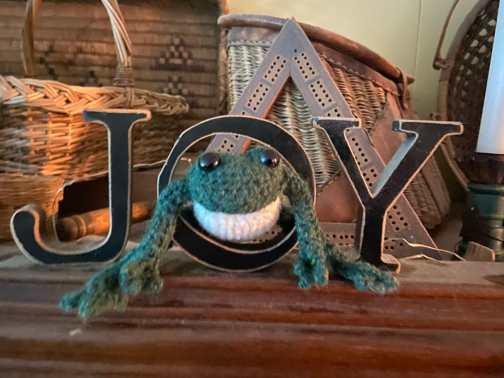 Image: small dark green frog with long bendable legs, black bead eyes, and a white belly on a wooden shelf. It is poking through a wooden letter “O” in the word “JOY”. Background is an old triangular crib board and woven baskets.