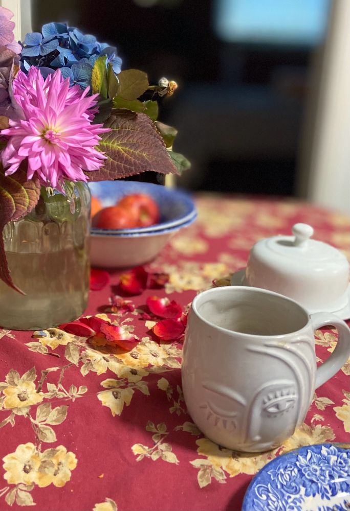 ID: photo of the final bouquet of flowers from my November garden (hydrangeas, dahlias, and a single rose) in a clear glass vase on the kitchen table, which is covered with a red, gold and olive flowered table cloth. The rose has lost all of its petals and they are on the table below the stem. Also visible: a white coffee cup, a covered butter dish, and the blurred edge of a blue dish of apples.
