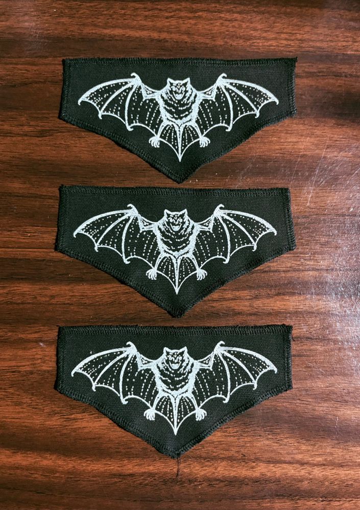 3 black patches with a bat design in white on a wood background 