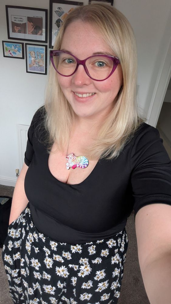 Selfie of a blonde lady with pink cat-eye glasses, wearing a black crop top and a long black skirt with a white daisies print all over it. Wearing a colourful tiger necklace!
