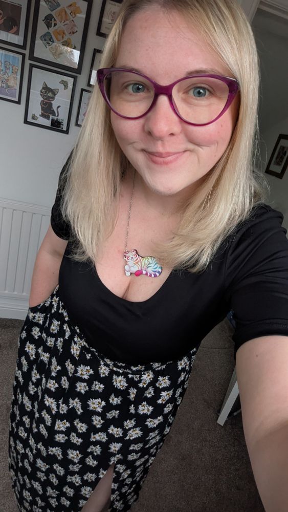 Selfie of a blonde lady with pink cat-eye glasses, wearing a black crop top and a long black skirt with a white daisies print all over it. Wearing a colourful tiger necklace!
