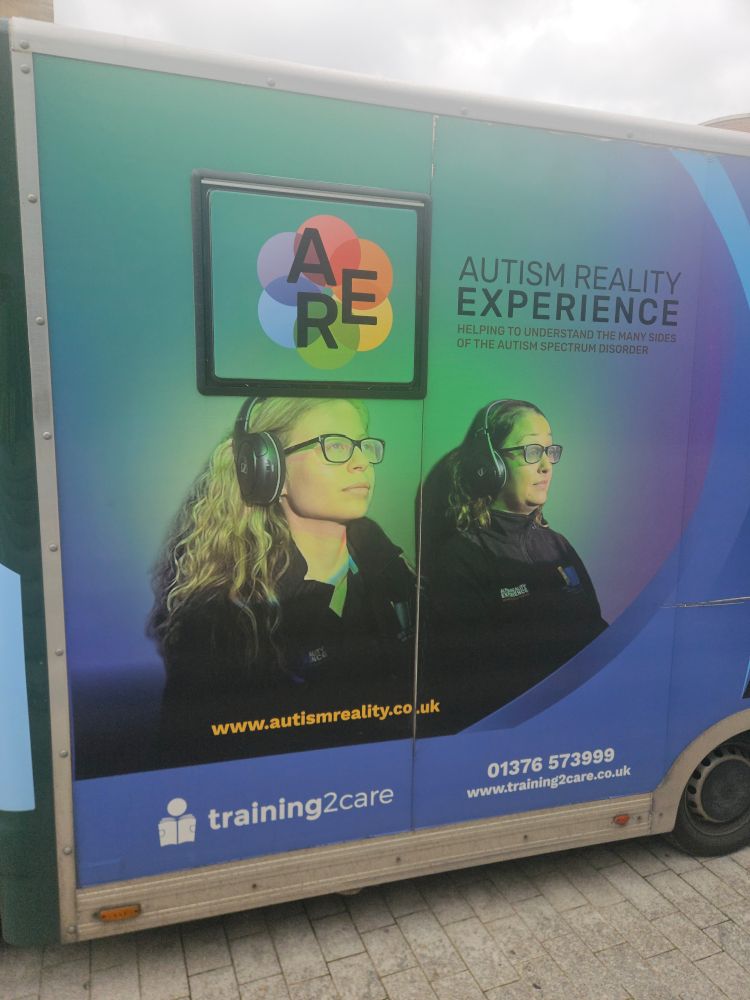 The side of a van advertising the Autism Reality Experience (A.R.E). In the advert two women with neutral expressions are sitting in chairs, wearing headphones under a green light.